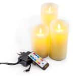 Drie Oplaadbare LED waxinekaarsen van 3 stuks DORSETTO staan met een afstandsbediening en voedingsadapter op een wit oppervlak. Deze kaarsen hebben een realistisch vlameffect en stralen een warmgeel licht uit.
