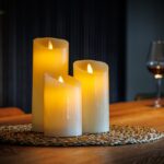 Drie Oplaadbare LED wax kaarsen van 3 stuks DORSETTO met oplichtende gele lampjes zitten op een geweven placemat op een houten tafel, terwijl een wijnglas en een fles rode wijn te zien zijn op de onscherpe achtergrond.