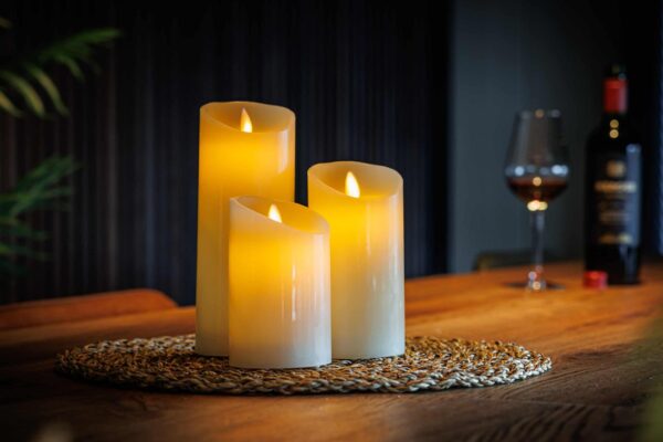 Drie oplaadbare LED wax kaarsen van 3 stuks DORSETTO in verschillende hoogtes verspreiden een warme gloed op een gevlochten mat op een houten tafel, terwijl op de achtergrond een wijnfles en gevuld glas zorgen voor een gezellige sfeer.