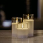 De DORSETTO LED kerzen set - oplaadbaar 105 uur met 3D vlam en afstandsbediening brandt sfeervol dichtbij elkaar in glazen houders op een egale ondergrond, tegen een zacht vervaagde donkere achtergrond.