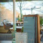 Op tafel: een LED dinerkaars 270mm oplaadbaar 130 uur warm wit (onderdeel van een 6-delige set met afstandsbediening), gloeiend in een glazen houder, naast een votief met patroon en een houten menustandaard. Buiten zijn vage ramen en bomen zichtbaar.