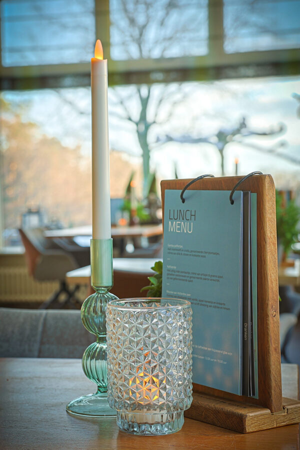 Op tafel: een LED dinerkaars 270mm oplaadbaar 130 uur warm wit (onderdeel van een 6-delige set met afstandsbediening), gloeiend in een glazen houder, naast een votief met patroon en een houten menustandaard. Buiten zijn vage ramen en bomen zichtbaar.