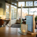 Een gezellige restauranttafel is voorzien van een Oplaadbare LED dinerkaars 270mm 130 uur warm wit (12 stuks, met afstandsbediening) in een glazen houder. Zonlicht stroomt door de grote ramen, waardoor de uitnodigende sfeer en het uitzicht op de stoelen en bomen buiten worden benadrukt.