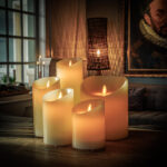 Een Oplaadbare LED wax kaars D 7,5 X 15 CM - warm wit gloeit op een houten tafel in een gezellige, zacht verlichte woonkamer, met een portretschilderij en geweven lamp op de achtergrond.
