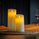 Twee gloeiende Oplaadbare LED wax kaarsen set van 2 DORSETTO zitten op een geweven placemat op een houten tafel, met een glas rode wijn en een fles op de zacht verlichte achtergrond, waardoor een uitnodigende en gezellige sfeer ontstaat.