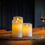 Een set van vier Oplaadbare LED wax kaarsen van DORSETTO gloeit warm op een geweven mat op een houten tafel, terwijl een wijnfles en glas op de achtergrond bijdragen aan de gezellige, uitnodigende sfeer.
