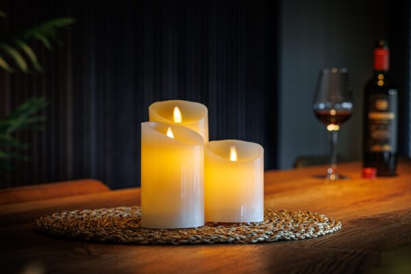 Een set van vier Oplaadbare LED wax kaarsen van DORSETTO gloeit warm op een geweven mat op een houten tafel, terwijl een wijnfles en glas op de achtergrond bijdragen aan de gezellige, uitnodigende sfeer.