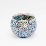 De Waxinelichthouder 9 CM Dorsetto mozaïek multicolor heeft een rond glazen ontwerp met blauwe en zilveren reflecterende tegels, die een brandende kaars vasthouden tegen een effen witte achtergrond.