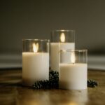 Drie DORSETTO led kaarsen uit de oplaadbare set (105 uur, 3D vlam, met afstandsbediening) branden in heldere glas houders op hout, omringd door groene sprietjes en creëren een warme, gezellige sfeer.