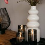 Een witte vaas met droogbloemen en de DORSETTO oplaadbare led kaarsen set 105 uur (3D vlam, afstandsbediening) staan op een ronde tray op hout. Op de achtergrond is deels een moderne zwarte draadlamp zichtbaar.
