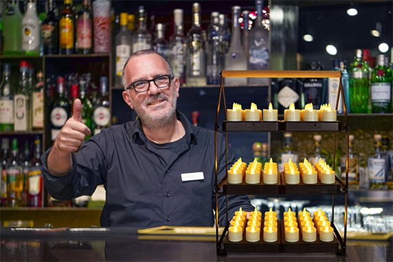 Een glimlachende barman met bril steekt zijn duim omhoog achter een met flessen beklede bar. Voor hem staat de Oplaadrek DORSETTO 35,5X21X51cm, een displaystandaard van drie niveaus met LED-kaarsen op batterijen.