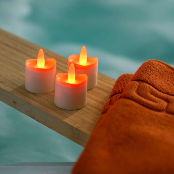 Zes RGB LED-Waxine theelichtjes (waterdicht, 68 uur oplaadbaar, met afstandsbediening) zitten op een houten plank naast een oranje handdoek, met een wazige achtergrond die een rustgevende spa of watersetting oproept.