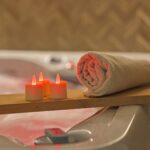 Een opgerolde beige handdoek en RGB LED-Waxine/theelichtjes waterdicht 68 uur oplaadbaar (6 stuks) met afstandsbediening gloeien op een houten plank boven een gevulde badkuip en creëren zo een ontspannen spa-achtige sfeer.