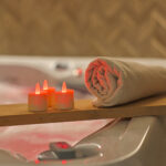 Een opgerolde beige handdoek en drie RGB LED-Waxine/theelichtjes oplaadbaar waterdicht (12 stuks) met afstandsbediening rusten op een houten plank boven een bad met roze getint water, waardoor een ontspannen spa-achtige sfeer wordt gecreëerd.