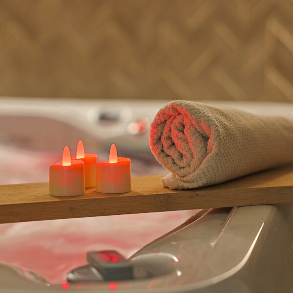 Een opgerolde beige handdoek en drie RGB LED-Waxine/theelichtjes oplaadbaar waterdicht (12 stuks) met afstandsbediening rusten op een houten plank boven een bad met roze getint water, waardoor een ontspannen spa-achtige sfeer wordt gecreëerd.