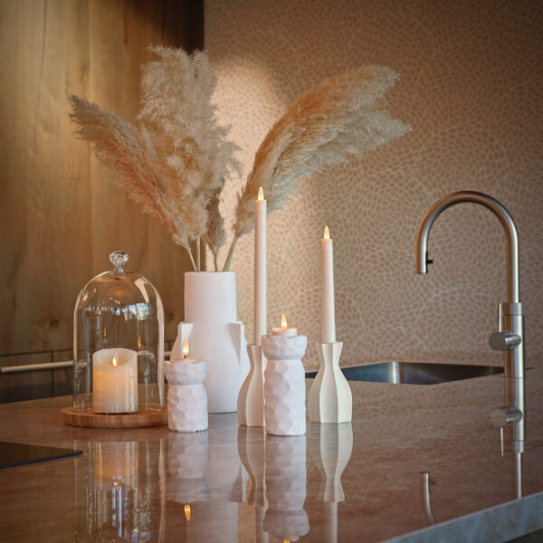 Een modern aanrecht met decoratieve witte vazen van pampagras, kaarsen in witte houders, een Oplaadbare LED dinerkaars 270mm (130 uur, warm wit, 12 stuks, met afstandsbediening), een kaars in glas en een strakke metalen kraan.