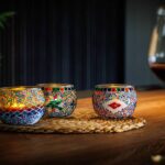 Drie mozaïek kandelaars van Theelichthouder Combie 9 CM Dorsetto, waarvan er een oplicht, staan op een geweven mat boven op een houten tafel. Rechts staat een glas rode wijn; zachte verlichting en een bladplant vormen de achtergrond.