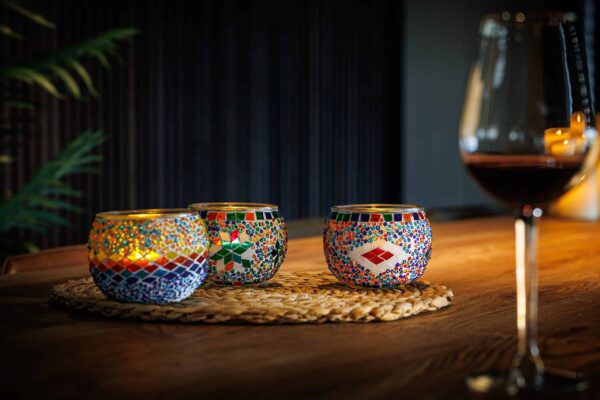 Drie mozaïek kandelaars van Theelichthouder Combie 9 CM Dorsetto, waarvan er een oplicht, staan op een geweven mat boven op een houten tafel. Rechts staat een glas rode wijn; zachte verlichting en een bladplant vormen de achtergrond.