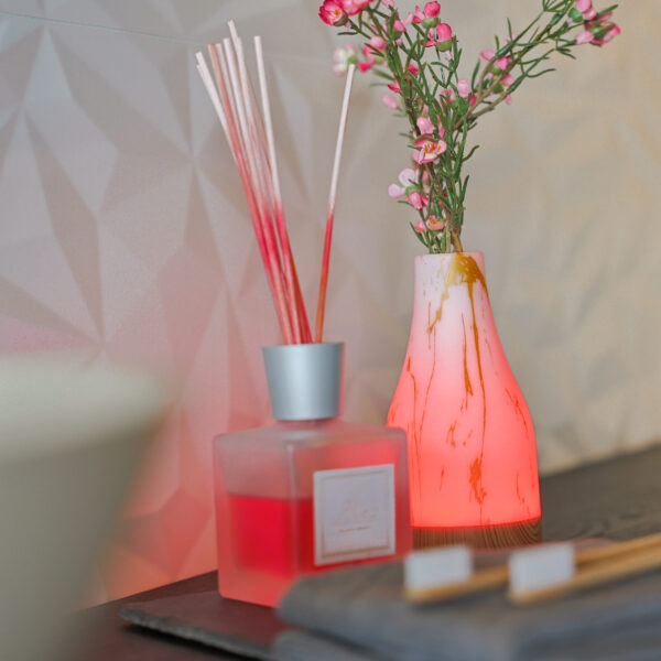 Een gematteerd glazen rietrooster met rode vloeistof staat naast een wit en roze gemarmerde vaas en de Tafellamp DORSETTO oplaadbaar 105 uur, ovaal, waterdicht, marmer groen houtlook grijs goud met afstandsbediening op een donker oppervlak tegen een muur met textuur.