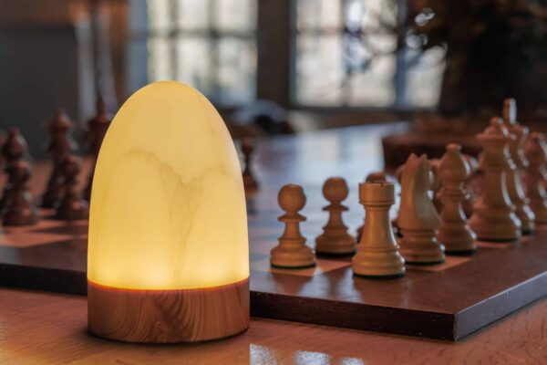 De oplaadbare tafellamp DORSETTO (105 uur, waterdicht, ovaal, marmer wit/grijs houtlook) met afstandsbediening staat op een tafel naast een schaakbord in een warm verlichte kamer met ramen op de achtergrond.