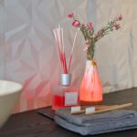 Een close-up van een aanrecht met twee tandenborstels, grijze handdoeken, een diffuser en de Tafellamp DORSETTO oplaadbaar 105 uur waterdicht ovaal marmer groen houtlook grijs goud met afstandsbediening die oplicht naast roze bloemen op een muur met structuur.