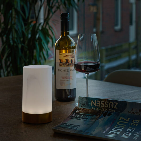 Een wijnfles, een gevuld glas en de Tafellamp DORSETTO oplaadbaar 105 uur (waterdicht, ovaal, marmer groen houtlook grijs goud, met afstandsbediening) gloeien op een houten tafel naast een tijdschrift en een groene plant bij een raam.