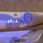 Een opgerolde handdoek en drie RGB LED-Waxine/theelichtjes oplaadbaar waterdicht (12 stuks) met afstandsbediening creëren een ontspannen, spa-achtige sfeer op een houten plank boven een bubbelbad.