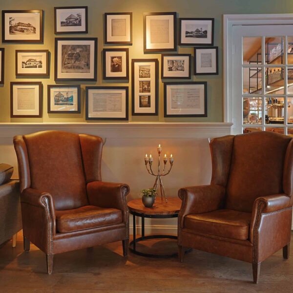 Twee bruine leren fauteuils staan naast een kleine ronde tafel met LED waxinelichtjes 3D (set van 12, oplaadbaar, timer, afstandsbediening, brandtijd tot 175 uur) en een plant. Ingelijste foto's en kunst bedekken de muur; een glazen deur onthult een warm verlichte kamer daarachter.