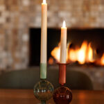 Twee kaarsen - een groene, een roodbruine - staan in glazen houders op een houten tafel. Daarachter gloeien een open haard en mozaïektegels warm op, aangevuld met een Oplaadbare LED dinerkaars 270mm (130 uur) met afstandsbediening voor een moderne touch.