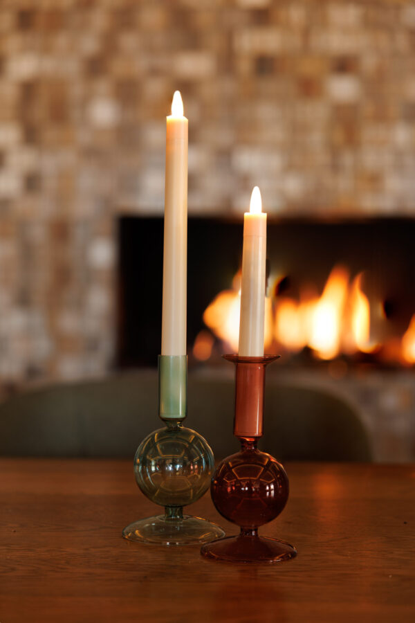 Twee kaarsen - een groene, een roodbruine - staan in glazen houders op een houten tafel. Daarachter gloeien een open haard en mozaïektegels warm op, aangevuld met een Oplaadbare LED dinerkaars 270mm (130 uur) met afstandsbediening voor een moderne touch.