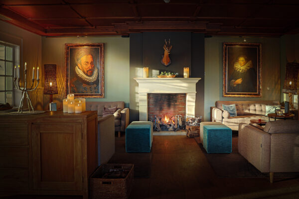 Een gezellige woonkamer met een verlichte open haard, sofa's, blauwe voetenbanken en klassieke portretten. Warme verlichting van LED dinerkaars oplaadbaar 97mm kaarsen (set van 6, warm wit, 130h met afstandsbediening) en een gemonteerde hertenkop boven de schouw creëren een gezellige sfeer.