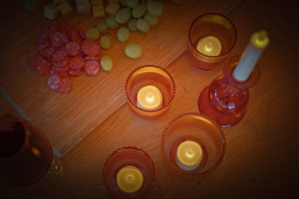 Een gezellig tafereel met Oplaadbare LED-was/theelichtjes (12 stuks, warm wit, 175 uur brandtijd, waterdicht, met afstandsbediening) in amberkleurige glazen houders naast een wijnglas en een houten plank met salami, kaas en druiven.