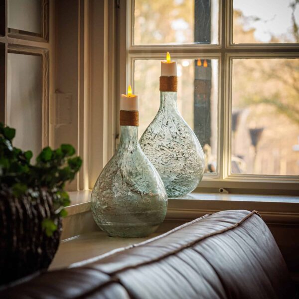 Twee getextureerde glazen vazen met 3D oplaadbare LED-thee/waxinelichtjes 175 uur - met Afstandsbediening - 12 stuks - timerfunctie erop staan op een vensterbank achter een bruine leren bank, bij een potplant, met bomen zichtbaar door het raam.