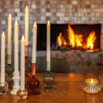 Een gezellig tafereel met brandende kaarsen in kleurrijke houders, bloemen in een vaas, een LED dinerkaars 270mm oplaadbaar 130 uur warm met 6 stuks met afstandsbediening op een houten tafel, en een gloeiend vuur in de open haard.