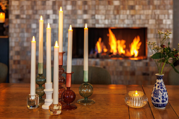 Een gezellig tafereel met brandende kaarsen in kleurrijke houders, bloemen in een vaas, een LED dinerkaars 270mm oplaadbaar 130 uur warm met 6 stuks met afstandsbediening op een houten tafel, en een gloeiend vuur in de open haard.