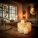 In een gezellige woonkamer gloeien de Oplaadbare LED waxinelichtjes D12,5 X H25 CM op een houten tafel, naast een kandelaar met kaarsen, een bank, een rieten lamp en een groot portret van een man. Warme sfeerverlichting vult de ruimte.