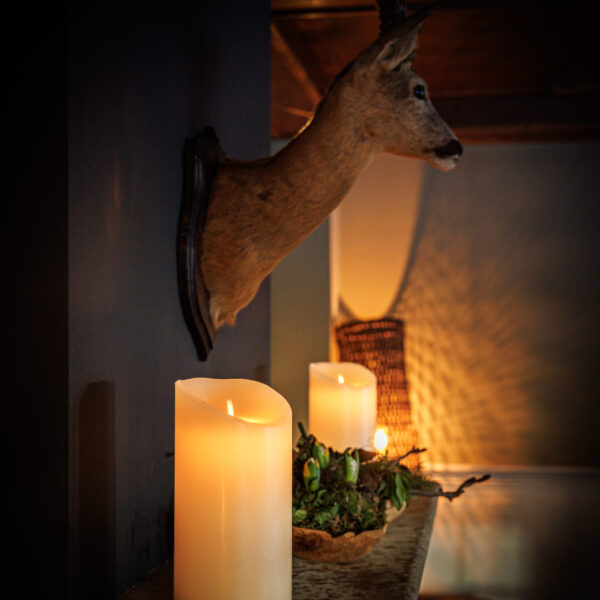 Een warm verlicht interieur heeft een gemonteerde hertenkop boven een schouw, versierd met de Oplaadbare LED wax kaars D 7,5 X 15 CM - warm wit en een groen middenstuk. Zachte schaduwen en gezellige verlichting maken de ruimte uitnodigend.