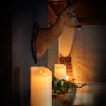 Een gemonteerde hertenkop hangt aan een donkere muur boven een richel versierd met een DORSETTO LED kleine was kaarsen set 2 kaarsen en rustiek groen, waardoor een gezellige, warme sfeer ontstaat.