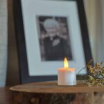 Een 3D oplaadbaar LED-thee/waxinelichtje (set van 12, 175 uur, met afstandsbediening en timerfunctie) geeft warm licht op een houten oppervlak naast bloemen, voor een ingelijste zwart-witfoto van een oudere vrouw.