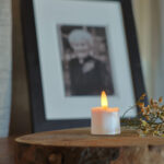 Een set 3D oplaadbare LED-thee/waxinelichtjes (175 uur, met afstandsbediening, timerfunctie, 6 stuks) gloeit tussen tere witte bloemen op hout voor een onscherp zwart-wit portret.