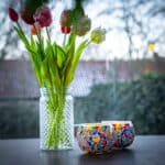 Een glazen vaas met kleurrijke kunsttulpen staat naast twee DORSETTO 9 cm donker groen theelichthouders op een tafel, tegen een zacht vervaagde buitenachtergrond.