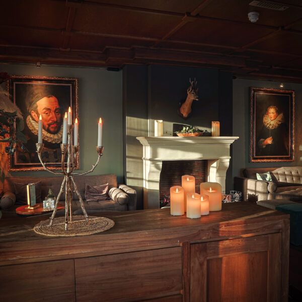 Een gezellige, elegante kamer met een brandende kandelaar en een Oplaadbare LED wax kaars D 7,5 X 15 CM - warm wit op een houten tafel, een sierlijke open haard met grote kaarsen, pluche banken, klassieke portretten en warme verlichting voor een vintage sfeer.