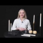 Een vrouw met lang blond haar zit aan een zwarte tafel versierd met witte kaarsen en de LED dinerkaars oplaadbaar 150 mm 58 uur warm wit (6 stuks met afstandsbediening). Ze draagt een helder wit overhemd tegen een zwarte achtergrond.