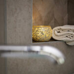 Een Theelichthouder zwart 9 CM DORSETTO staat op een badkamerplankje naast netjes opgevouwen crèmekleurige handdoeken, met een vage kraan op de voorgrond.