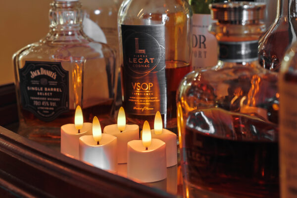 Een close-up van drankflessen, waaronder Jack Daniels en VSOP cognac, op een houten oppervlak met zes 3D oplaadbare LED-thee/waxinelichtjes (175 uur, met afstandsbediening en timerfunctie) die ervoor oplichten.