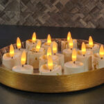 Een rond gouden dienblad bevat 6 oplaadbare LED waxinelichtjes 3D kaarsen (brandtijd 175 uur, met afstandsbediening en timer), die allemaal warm gloeien op een donker aanrecht tegen een betegelde muur met textuur.