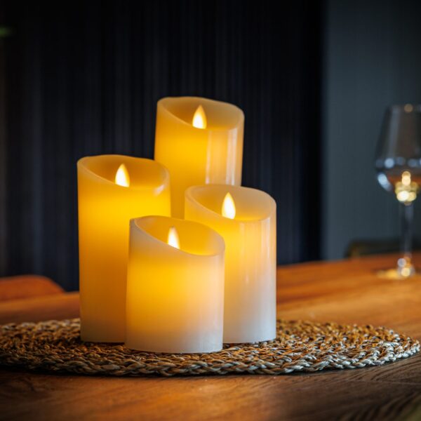 Een set van vier Oplaadbare LED wax kaarsen van 4 DORSETTO met verschillende hoogtes ligt op een geweven mat boven op een houten tafel en verlicht zacht de achtergrond waar een wijnglas en een fles de warme sfeer versterken.