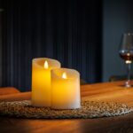 De LED kleine wax kaarsen set 2 kaarsen DORSETTO wordt weergegeven op een geweven mat op een houten tafel, met een glas rode wijn en een wijnfles op de achtergrond voor een warme, gezellige vibe.