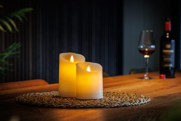 De LED kleine wax kaarsen set 2 kaarsen DORSETTO wordt weergegeven op een geweven mat op een houten tafel, met een glas rode wijn en een wijnfles op de achtergrond voor een warme, gezellige vibe.