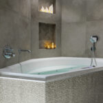 Een modern bad met een buitenkant van mozaïektegels staat tegen grijs betegelde muren. Twee verzonken planken erboven bevatten Oplaadbare LED waxine/theelichtjes 175 uur waterproof warm wit (6 stuks) met afstandsbediening en huisfiguurtjes, waardoor een gezellige sfeer wordt gecreëerd.